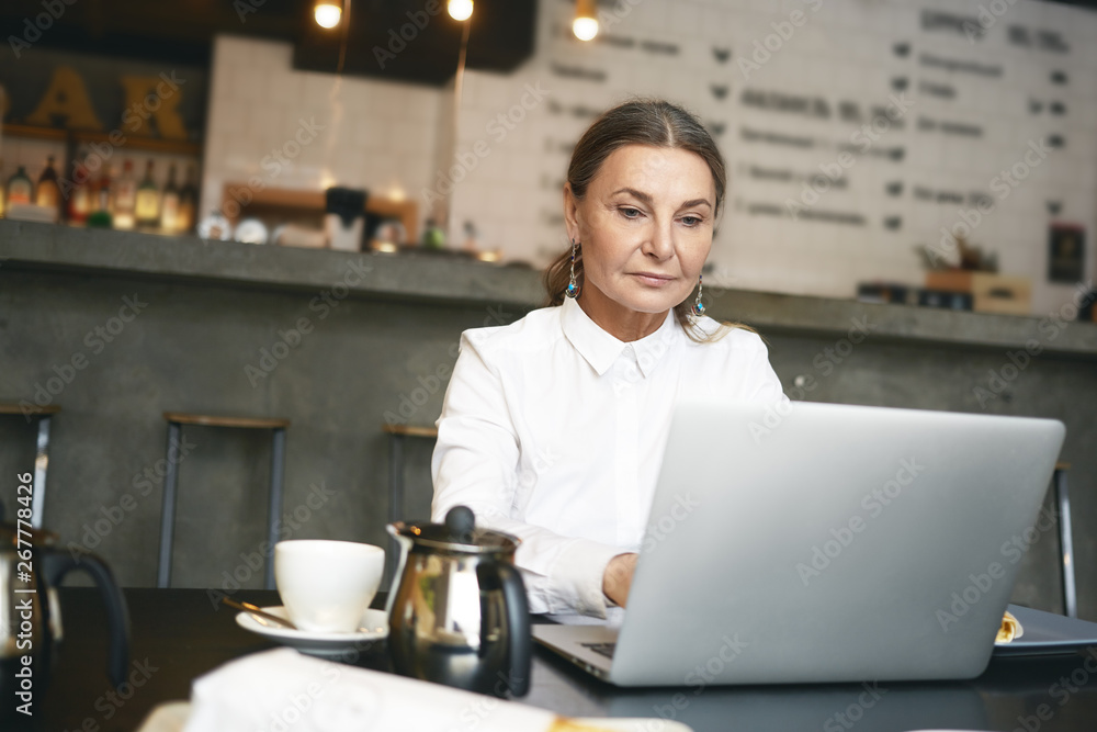 Picture of attractive middle aged businesswoman in formal wear browsing website for job search and staff recruitment on generic portable computer during break at coffee shop, having concentrated look Picture of attractive middle aged businesswoman in formal wear browsing website for job search and staff recruitment on generic portable computer during break at coffee shop, having concentrated look