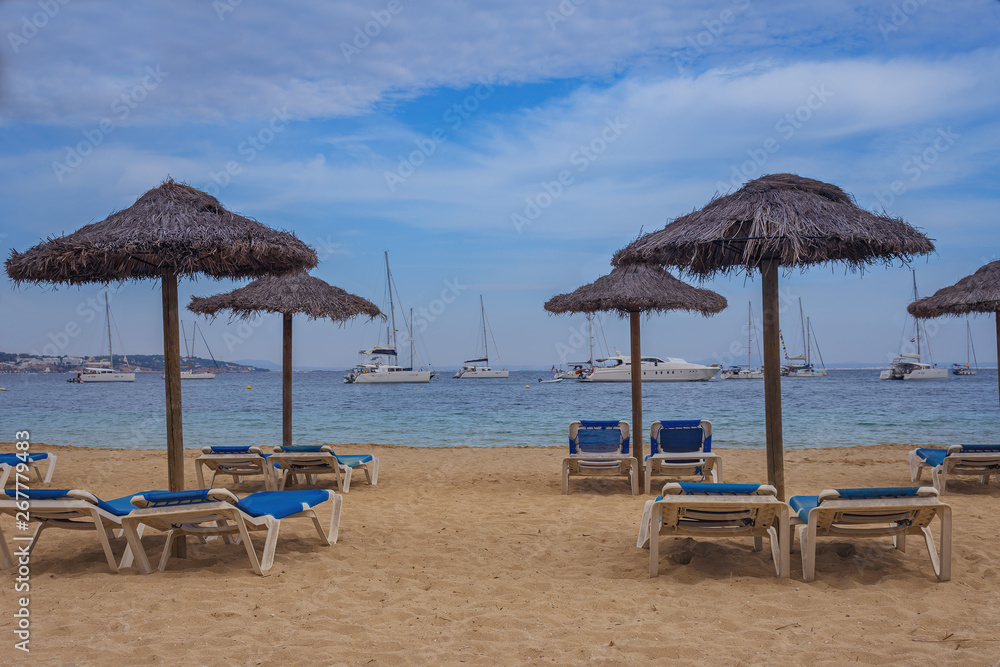 sun loungers and straw umbrellas on the sandy beach  of the Spanish town of Magaluf against the blue sea and white yachts