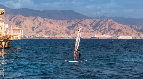 a child windsurfer sailing into the eilat marina in israel with the port of akaba jordan in the background
