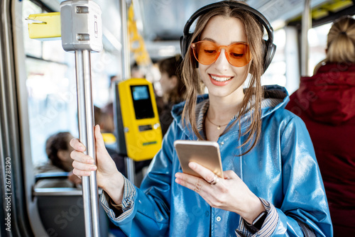Young stylish woman using public transport, standing with headphones and smartphone while moving in the modern tram