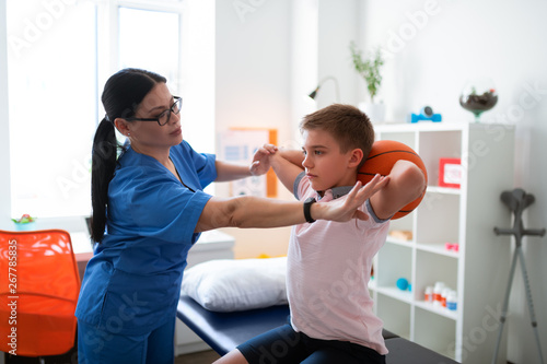 Gentle dark-haired Asian doctor correcting posture of little kid photo