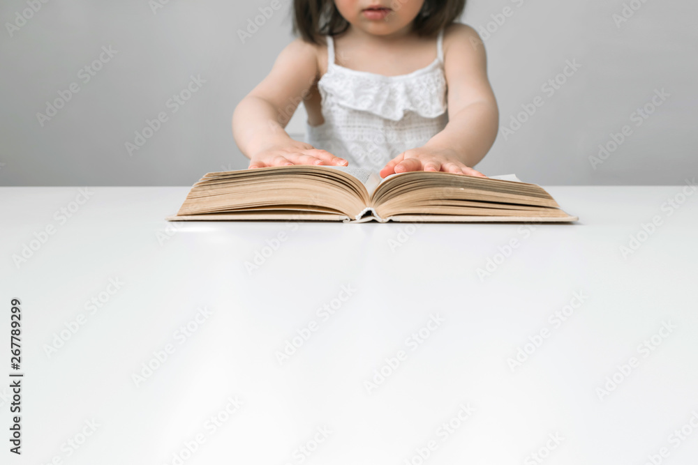 The little girl opened the book. The child is studying the book. A girl is reading a book. The book is on a white table. The girl in white overalls. Girl with a short haircut The little girl opened the book. The child is studying the book. A girl is reading a book. The book is on a white table. The girl in white overalls. Girl with a short haircut