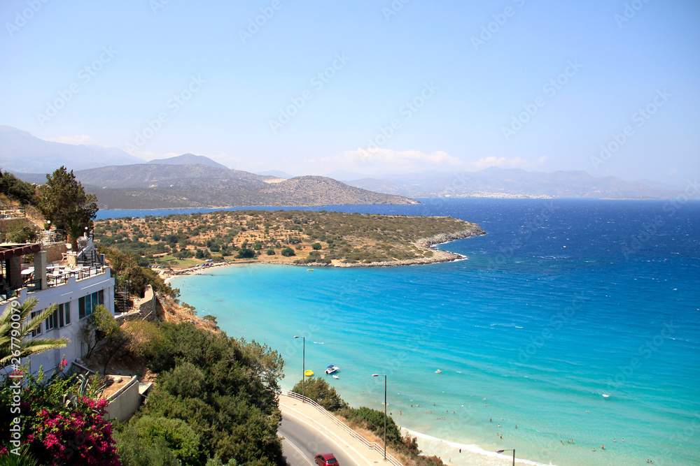 An island coast with clear blue sea, trees above the water by sandy beach and hills on a summer day, Mediterranean sea in turquoise lagoon