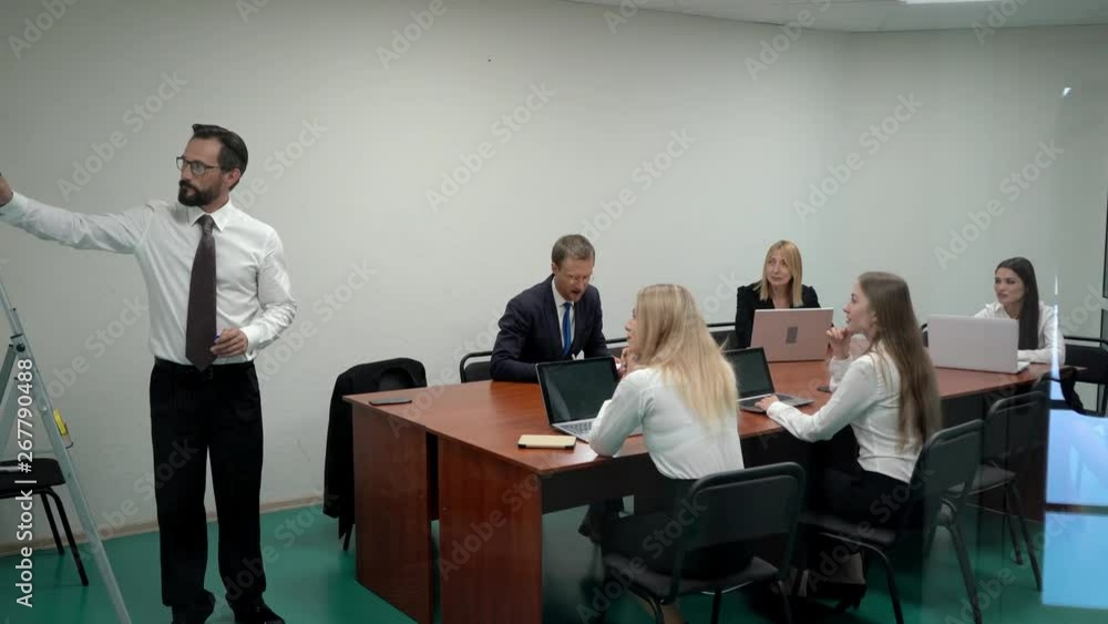 A businessman discussing a flip chart on a white board in a meeting room to working team while standing in the office. A businessman discussing a flip chart on a white board in a meeting room to working team while standing in the office.
