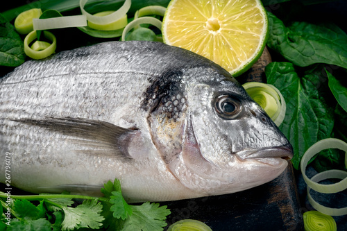 Fresh and raw sea bream fish with lemon and herbs on a black wooden background, top view and close up