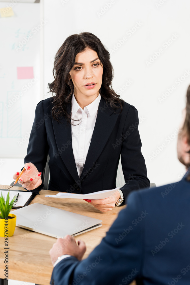selective focus of brunette recruiter holding paper and pencil and looking at man selective focus of brunette recruiter holding paper and pencil and looking at man
