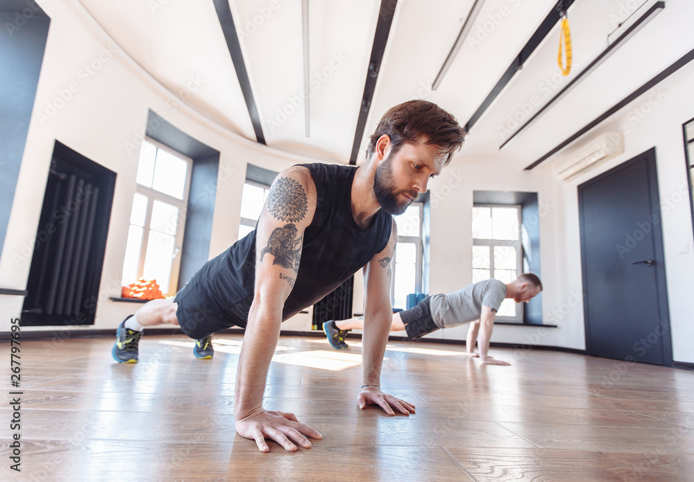 Two friends men in good physical shape doing workout together in the gym. Concept of team spirit and joint visits to the hall for support. Side view of two young sportman in sportswear doing plank
