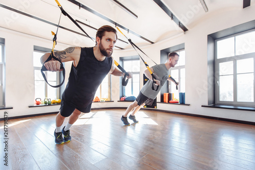 Two fit strong athletic male friends are training together with trx in the gym. Concept of strength training and resistance exercises