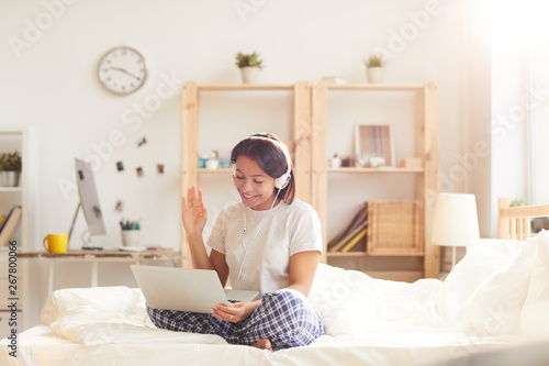 Portrait of smiling Mixed-Race woman using video chat sitting on bed lit by sunlight, copy space