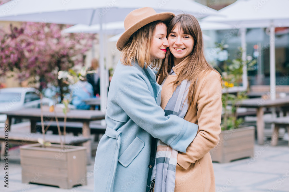 Fototapeta premium Best girlfriends hug kiss and love.. Adult emotional women sisters dressed in stylish beige and blie coat, brown hat. Hipster. Warm toning. The concept of true friendship.