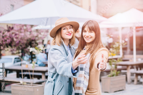 Best girlfriends hugпштп gesturing beckoning you at camera. Adult emotional women sisters dressed in stylish beige and blie coat, brown hat. The concept of true friendship. Sun flare effect