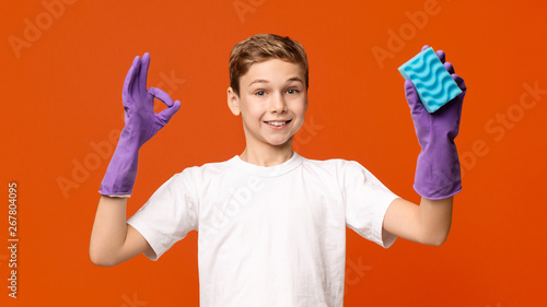 Cute boy in cleaning gloves showing OK sign and holding sponge