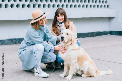 Two stylish women plays with a dog at city street urbam background. Training Golden retriever, the performance of the teams. photo