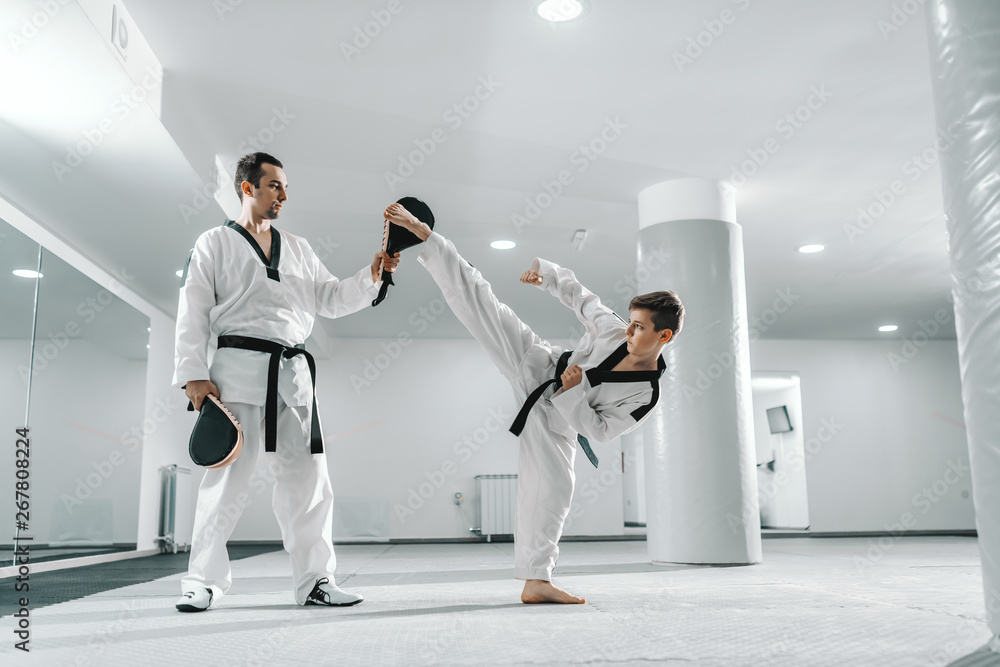 Fototapeta Young Caucasian boy in dobok kicking barefoot while trainer holding kick target. Taekwondo training concept.