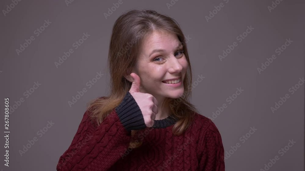 Pretty young female student puts a thumb up smilingly into camera to show like and respect on gray background. Pretty young female student puts a thumb up smilingly into camera to show like and respect on gray background.