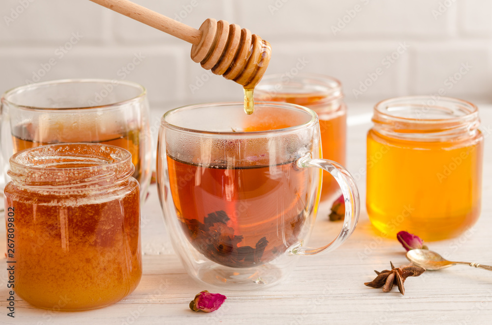 Polyfloral honey flows from a wooden honey spoon. A viscous stream of sweet amber flower honey flows from a wooden honey spoon into a glass cup of tea. Polyfloral honey flows from a wooden honey spoon. A viscous stream of sweet amber flower honey flows from a wooden honey spoon into a glass cup of tea.