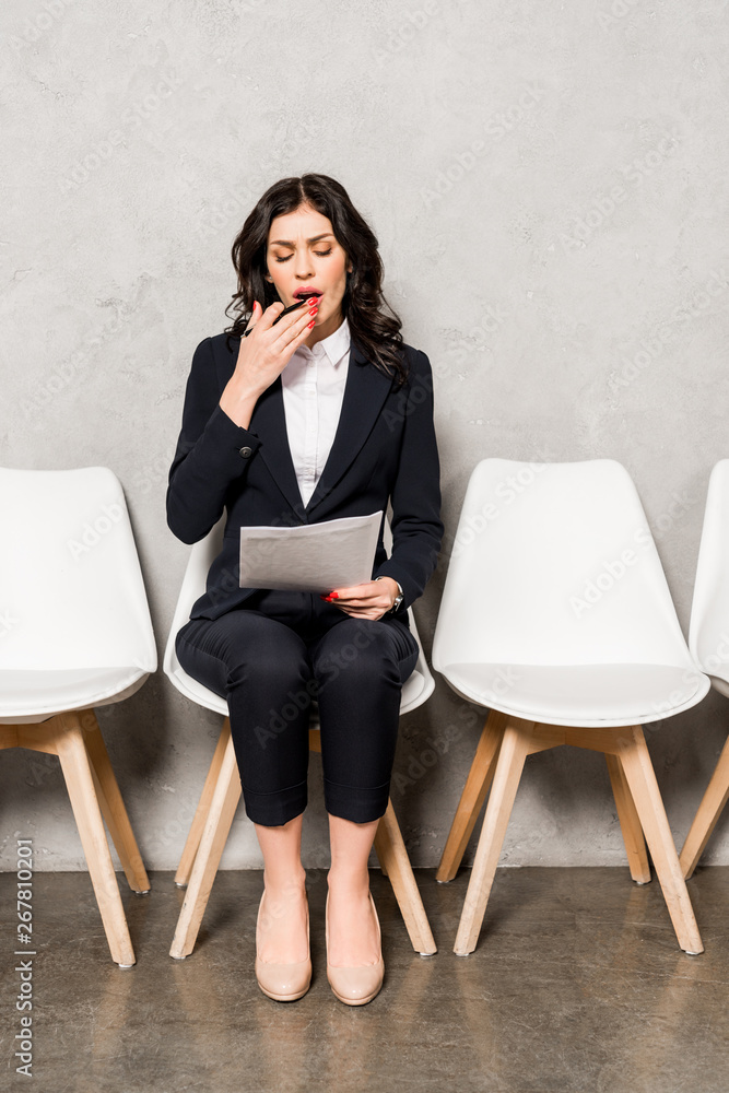 tired brunette woman looking at paper and yawning while sitting on chair tired brunette woman looking at paper and yawning while sitting on chair