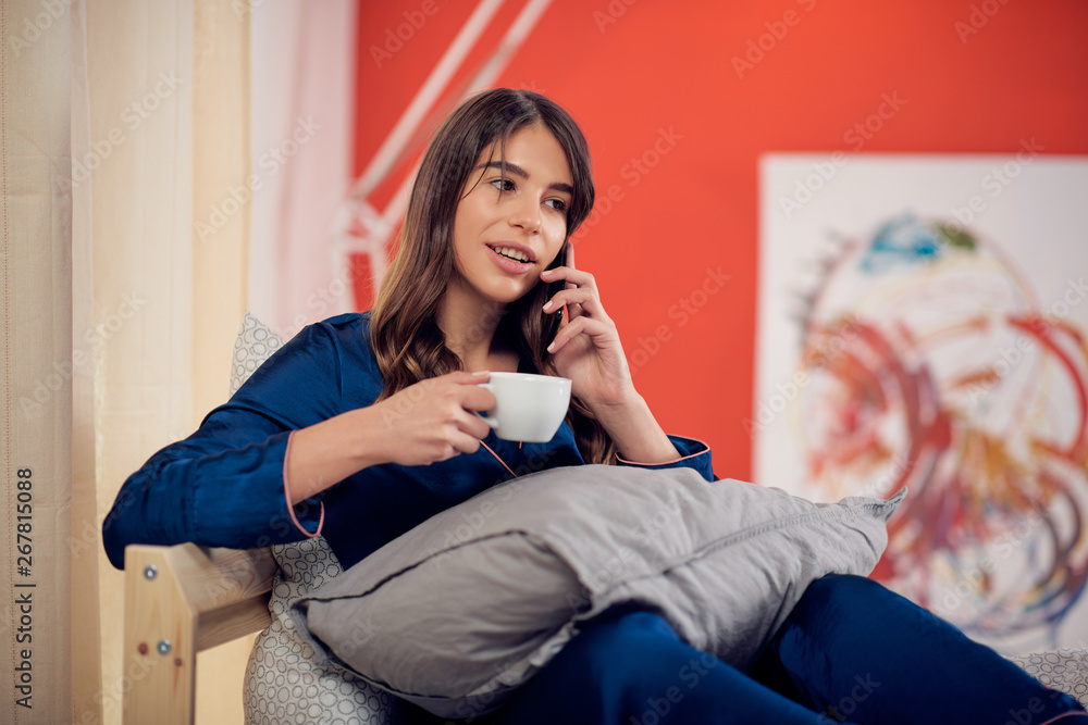Beautiful Caucasian brunette in pajamas sitting on the bed in bedroom, talking on the phone and drinking her morning coffee.