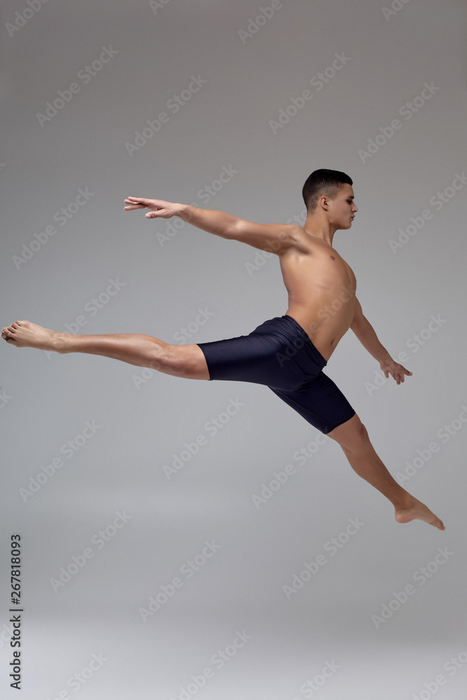 Fototapeta premium Photo of a handsome man ballet dancer, dressed in a black shorts, making a dance element against a gray background in studio.