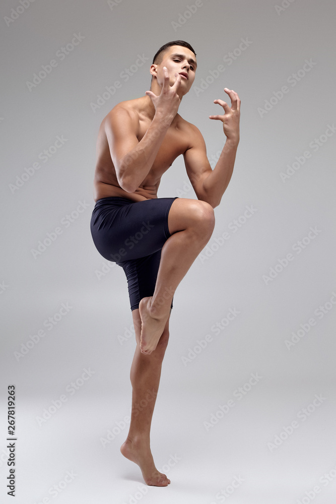 Obraz premium Photo of a handsome man ballet dancer, dressed in a black shorts, making a dance element against a gray background in studio.