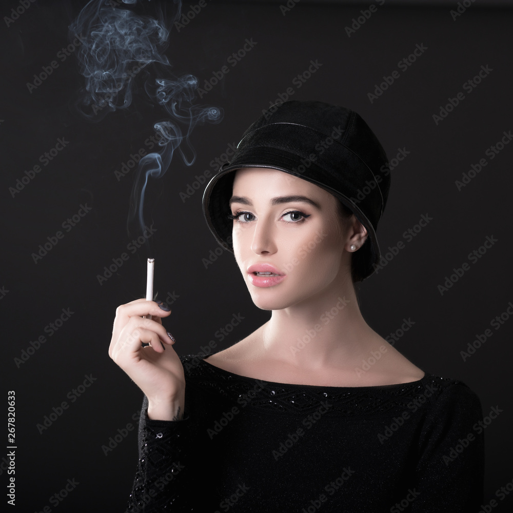 Obraz premium Young fashion woman smoking cigarette in hat and black drees over dark background. Vintage female portrait, styling. Image toned.