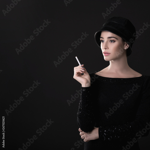 Young fashion woman smoking cigarette in hat and black drees over dark background. Vintage female portrait, styling. Image toned.