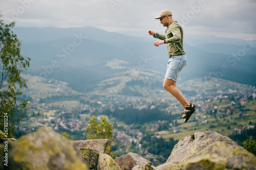 Lifestyle summer portrait of sportive man jumping from stone on top of mountaing with beautiful landscape in front. Male traveler having fun among hils and rocks at wild nature outdoor.