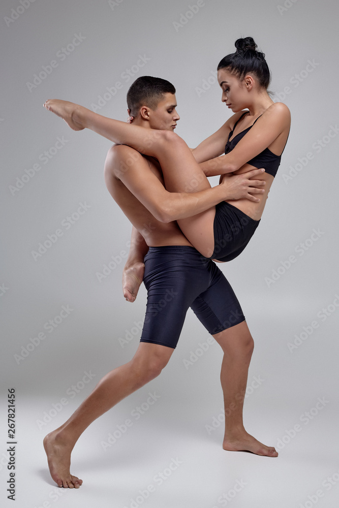The couple of a young modern ballet dancers in black suits are posing over a gray studio background. The couple of a young modern ballet dancers in black suits are posing over a gray studio background.
