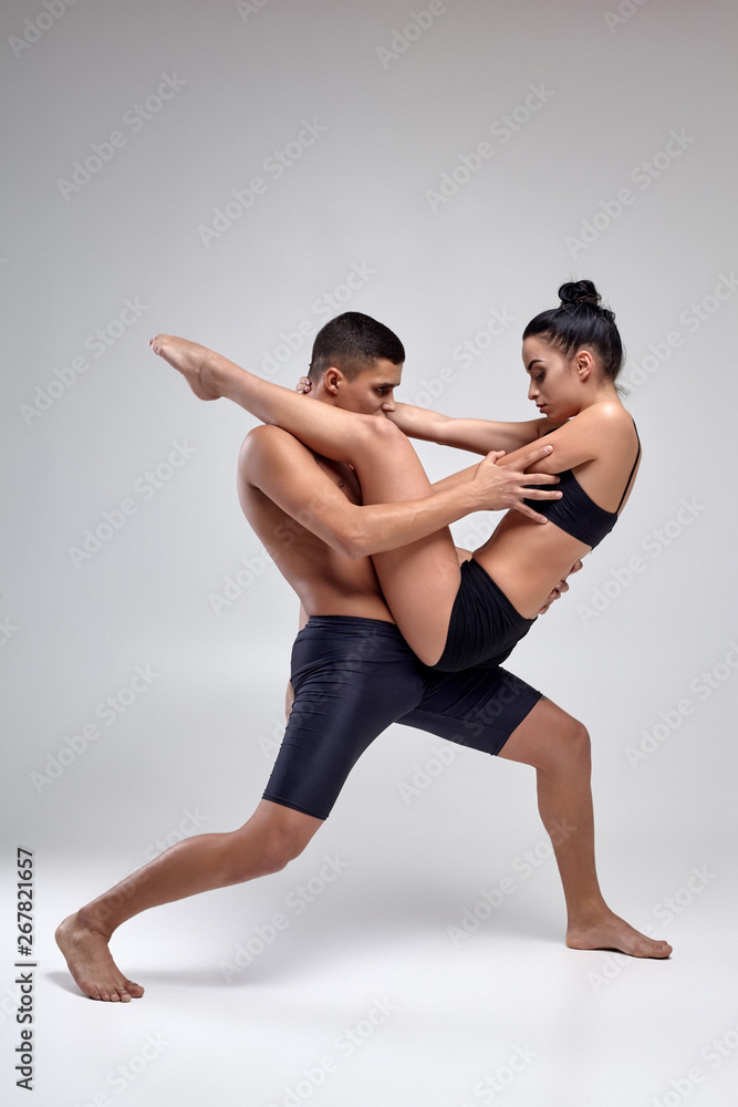 The couple of a young modern ballet dancers in black suits are posing over a gray studio background. The couple of a young modern ballet dancers in black suits are posing over a gray studio background.