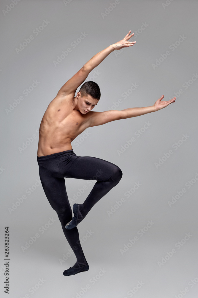 Photo of an athletic man ballet dancer, dressed in a black tights and pointe, making a dance element against a gray background in studio.