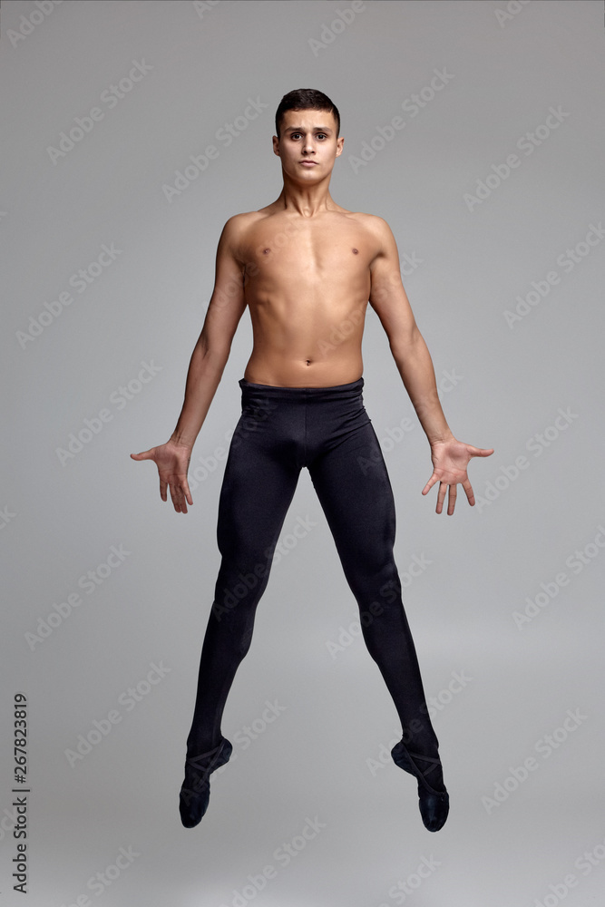 Obraz premium Photo of an athletic man ballet dancer, dressed in a black tights and pointe, making a dance element against a gray background in studio.