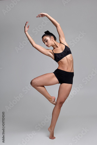 Studio photo of a beautiful woman ballerina dressed in black two-piece swimsuit doing a dance element on a gray background.