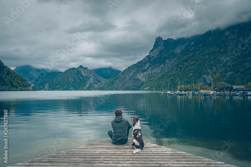 Mężczyzna z psem siedzą na pomoście nad jeziorem Traunsee w Austrii w górach w pochmurny dzień.