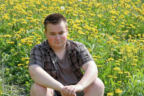 A young plump man sits in a meadow with blooming dandelions. Pensively looking in front of him.