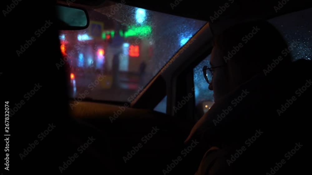 The girl in round glasses sits in the passenger seat in a dark car and looks through the windshield. Outside multicolored reflections in a blurred window with water drops. Rainy night weather. The girl in round glasses sits in the passenger seat in a dark car and looks through the windshield. Outside multicolored reflections in a blurred window with water drops. Rainy night weather.