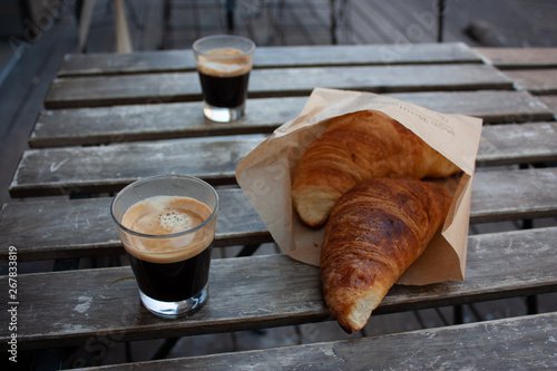 Zbliżenie na dwa kieliszki espresso i croissanty, stojące na drewnianym rustykalnym stoliku na zewnątrz kawiarni