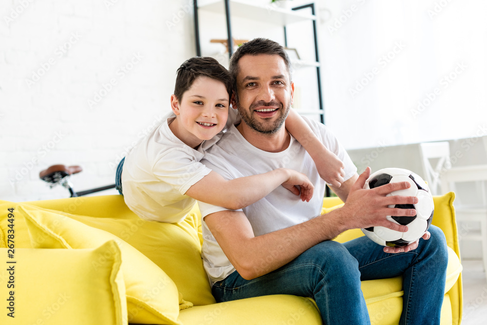 happy father and son on couch with soccer ball looking at camera at home happy father and son on couch with soccer ball looking at camera at home