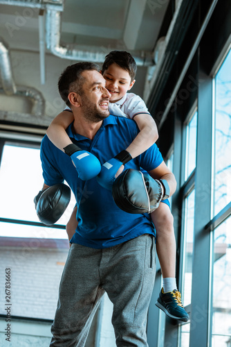 happy father giving piggyback ride to smiling son in boxing gloves at gym