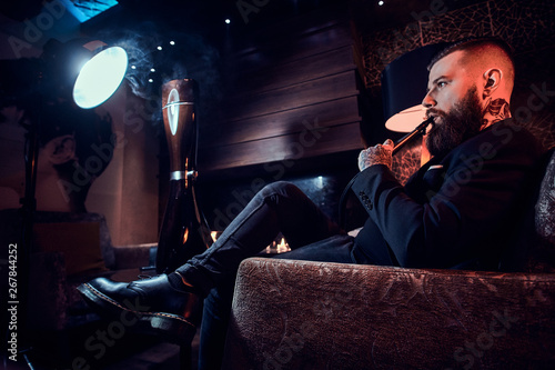 Handsome tattooed man is relaxing on the armchair and is smoking hookah.