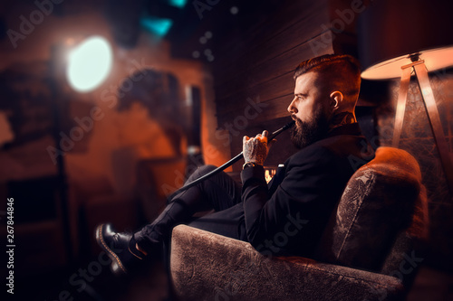 Handsome bearded man is relaxing on lounge while smoking hookah. He has tattoo on his hand.