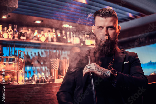Portrait of tattooed bearded man which is smoking hookah, making nice vapour while sitting near bar counter
