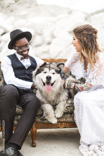 Best friend animals concept - Caucasian woman and African man touch head to head his beautiful tender dog in outdoor desert location place sitting on a retro sofa © sofiko14