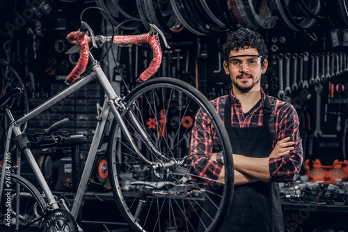 Smiling happy man in glasses is standing near fixed bicycle at his own workshop.