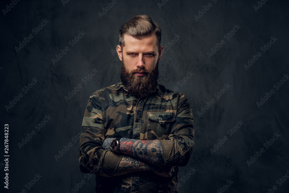A stylish bearded guy with tattooed hands in the military shirt, posing with his arms crossed and looking sideways. Studio photo against a dark wall A stylish bearded guy with tattooed hands in the military shirt, posing with his arms crossed and looking sideways. Studio photo against a dark wall