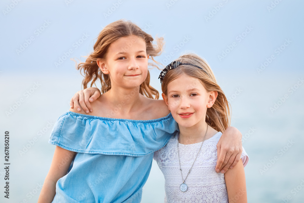 Two young girls wearing casual summer clothing looking, smiling at camera in a coastal region