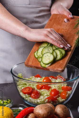 A young woman in a gray apron adds a cucumber to a salad