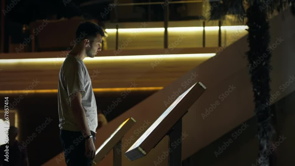 Attractive man using interactive touchscreen terminal display for searching info. Modern teen in wireless earphones. Evening time, lowlight exterior. Education and entertainment concept. Close up shot