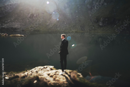 Full length portrait of a handsome young caucasian man dressed in suit sitting on a rock in the mountains near a lake against sunrise looking away.