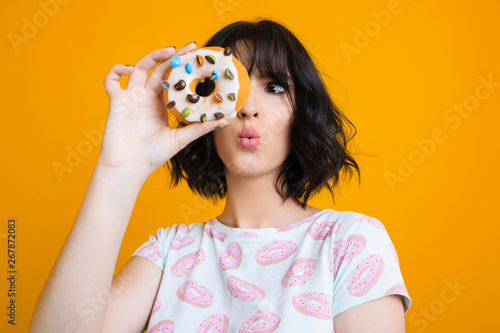 Beautiful adult female dressed in a shirt with donuts looking astonished through a donut in front of a yellow studio wall.