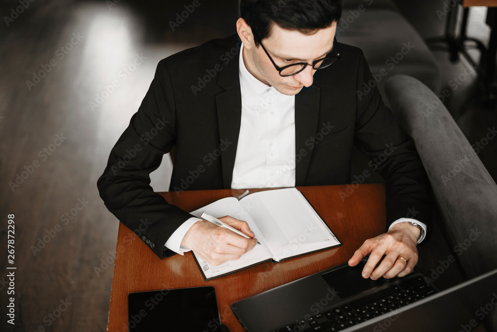 Upper view of a male hand writing in a notebook and operating on a laptop while sitting on a table. Upper view of a male hand writing in a notebook and operating on a laptop while sitting on a table.